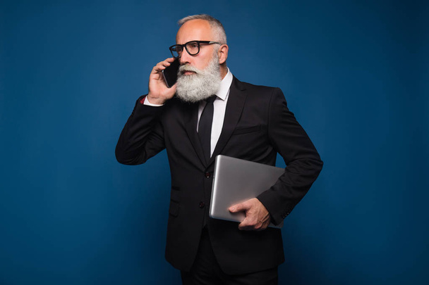 Elfoglalt szakállas súlyos és az üzletember tartózkodás-val jegyzetfüzet és mozgatható telefon elszigetelt kék háttérrel. Működő üzleti ember beszélni a telefon, egyenes öltönyt és szemüveg. - Fotó, kép