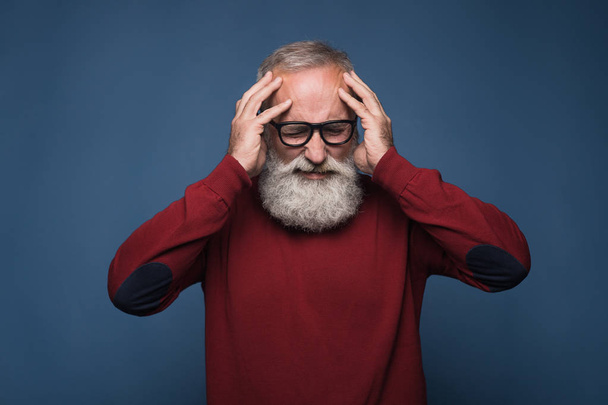 Kopfschmerzen. alter bärtiger Mann in rotem Sweater und schwarzer Brille zeigt Kopfschmerzen. Schmerzgefühle isolieren sich auf blauem Hintergrund. Stress nach einem anstrengenden Bürotag - Foto, Bild