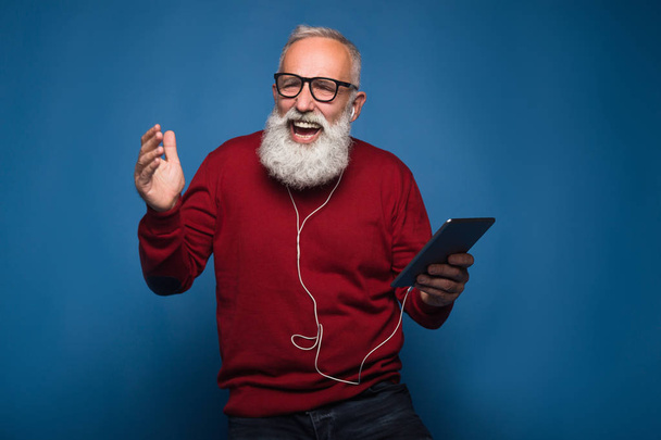 Verwachtingen van een nieuw lied. Gelukkig moderne bebaarde man luisteren een nieuwe muziek van Tablet PC. Man met hoofdtelefoon luisteren naar muziek geïsoleerd op blauwe achtergrond. Gelukkig dansen uit coole muziek op koptelefoon. - Foto, afbeelding