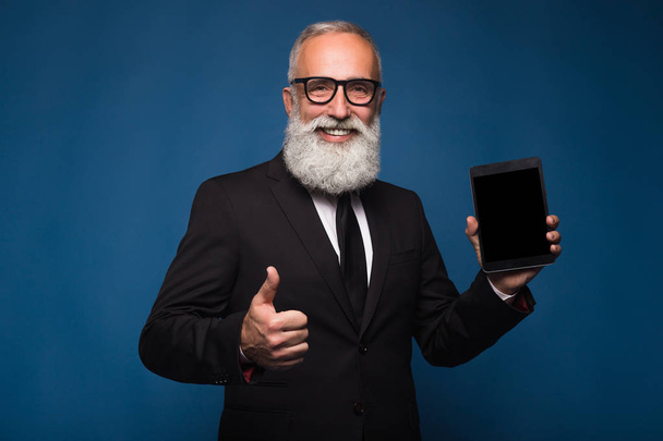 Felice uomo barbuto in abito formale tiene tablet digitale e mostra spazio copia sullo schermo mentre in piedi su sfondo blu. Posto vuoto di schermo di targa. Mostra uomo con lo schermo di tablet e pollice in su
 - Foto, immagini