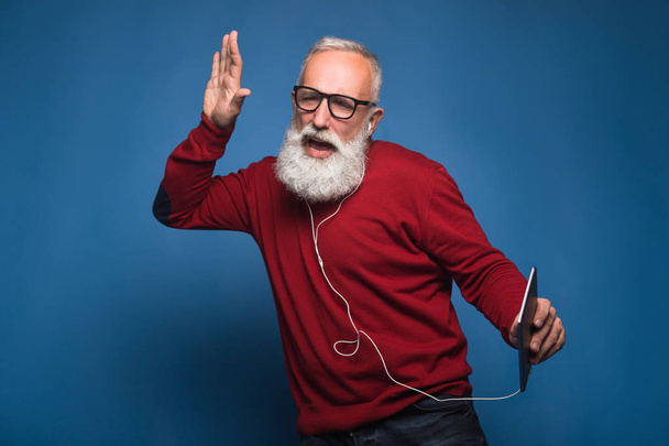 Очікування від нової пісні. Щасливий сучасний бородатий чоловік слухає нову музику з планшета. Чоловік з навушниками слухає музику ізольовано на синьому фоні. Щасливо танцює з крутої музики на навушниках
. - Фото, зображення