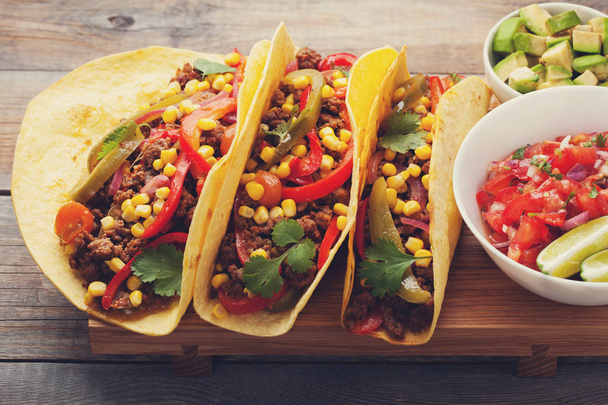 Kolme meksikolaista tacoa jauhelihalla ja sekoita vihanneksia vanhalla maalaispöydällä. Meksikolainen ruokalaji kastikkeella salsaa kulhossa ja avokado. Mausteista ja kuumaa katuruokaa. lähikuva
 - Valokuva, kuva