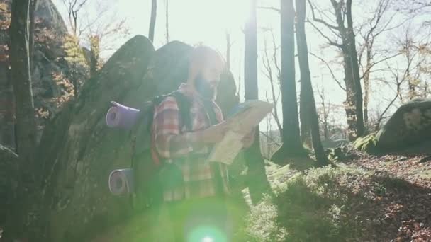 Stylish hipster man traveler with backpack holding map and exploring on top of mountains, travel concept. Autumn. A handsoome man wearing a beard keeps a tourist map in his hands - Materiał filmowy, wideo