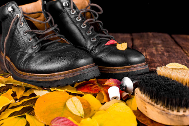 Impermeabile stivali neri su sfondo di legno con attrezzature di lucidatura foglie autunno, spazzola e crema lucida
. - Foto, immagini