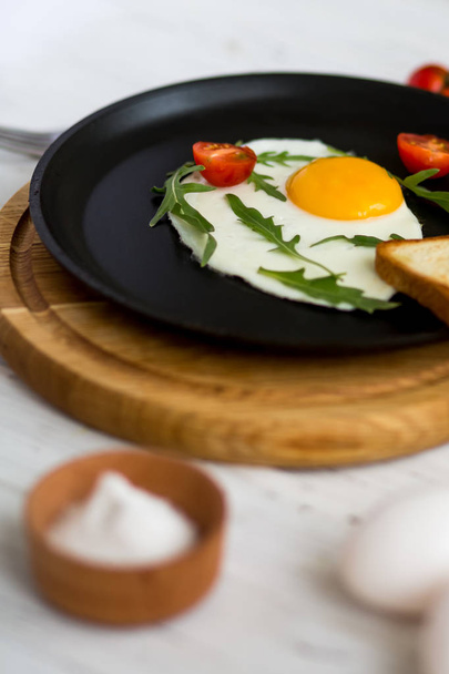 Chicken fried egg in a black frying pan with cherry tomatoes, arugula, salt. Breakfast on a white wooden table.  - Foto, Imagen
