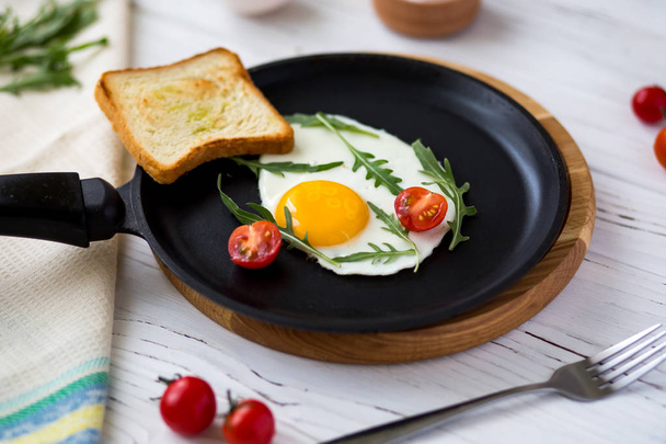 Kip gebakken ei in een zwarte koekenpan met cherrytomaatjes, rucola, zout. Ontbijt op een witte houten tafel.  - Foto, afbeelding