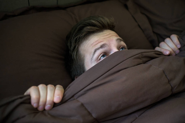 Καυκάσιος νεαρός άνδρας που κρύβονται στο κρεβάτι κάτω από την κουβέρτα στο σπίτι. - Φωτογραφία, εικόνα