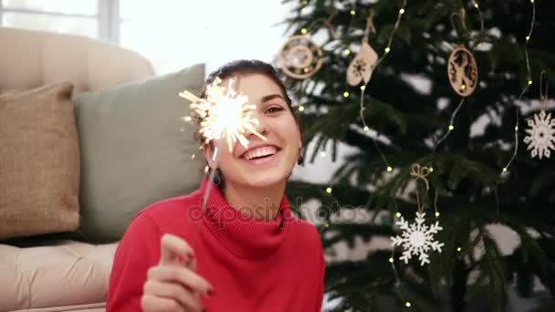 Smiling friendly attractive young woman sitting on the floor by the Christmas tree celebrating Xmas with sparklers and looking in the camera - Filmati, video