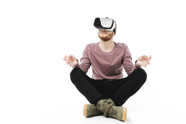 Young man sitting in studio and meditating while using virtual reality glasses isolated. Smiling boy playing with visual reality glasses on white background - Photo, Image