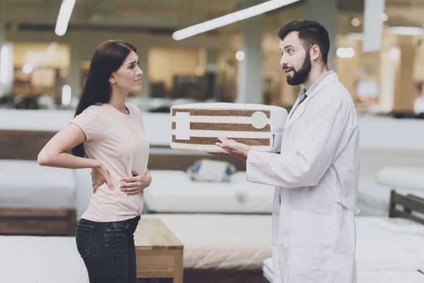 Consultant-orthopedist helps the girl to choose a mattress in the store. He offers her a mattress and shows his sample - Φωτογραφία, εικόνα