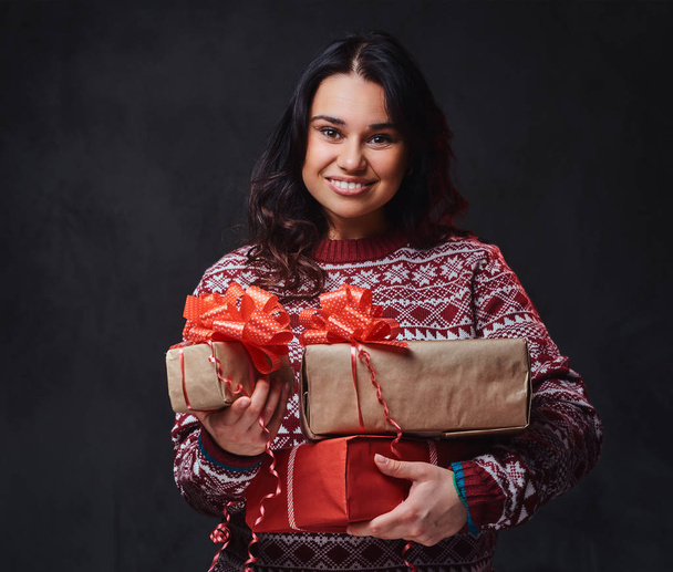 Πορτρέτο της εορταστική χαμογελώντας μελαχρινή γυναίκα με μακριά σγουρά μαλλιά, ντυμένη με ένα κόκκινο πουλόβερ κατέχει τα δώρα Χριστουγέννων. - Φωτογραφία, εικόνα
