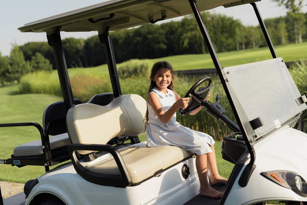 A little girl is sitting barefoot in a white golf car and hanging behind the wheel - Foto, imagen