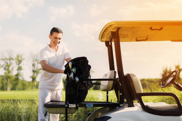 A man in white clothes stacks a bag with golf clubs on the trunk of a golf cart - Foto, imagen