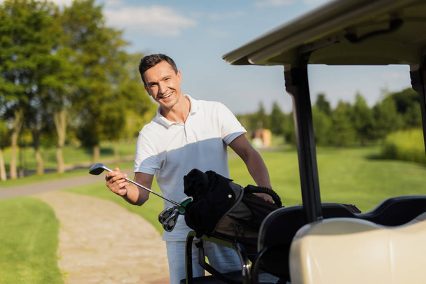 A smiling man pulls out of a bag with sticks, a golf club. Bag lies on the luggage compartment of the golf cart - Foto, Imagem