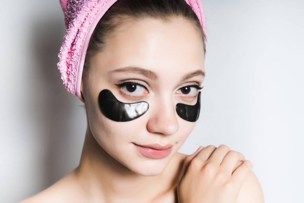 beautiful young girl looks after the skin of the face, under the eyes of black silicone patches, on the head a pink towel - Foto, afbeelding