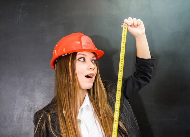 young surprised girl builder in a protective orange helmet holds a measuring tape in hand - Φωτογραφία, εικόνα