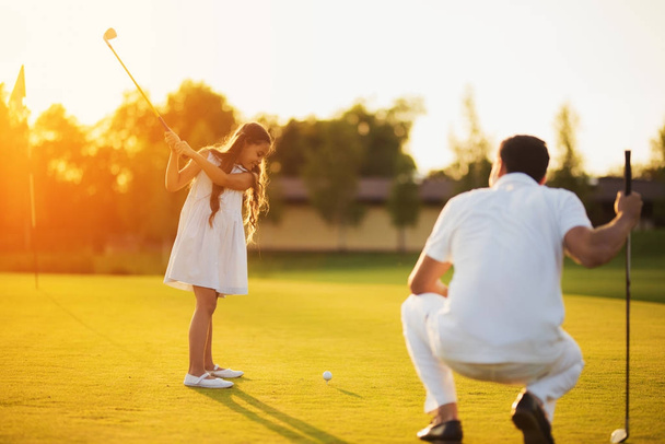 A man is squatting on a golf club and looking at a girl who is swinging a club to strike the ball - Foto, imagen