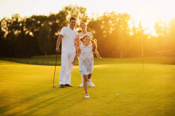 Happy family posing on a golf course on a sunset background. The girl smiles and runs towards the camera - 写真・画像