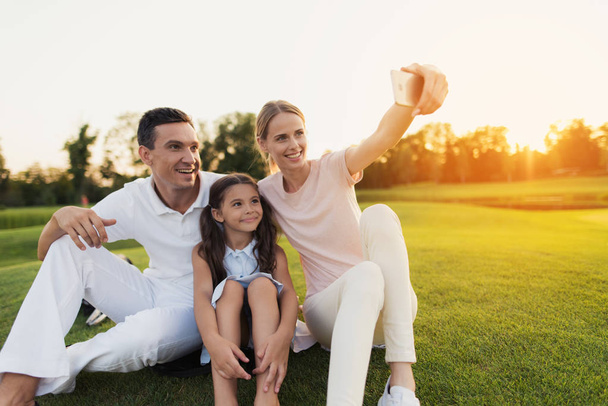 Familie doen selfie op grijze smartphone zittend op gras op golfbaan op zonsondergang achtergrond - Foto, afbeelding