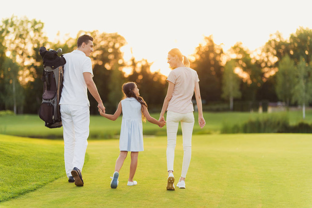 The family is walking along the golf course. Mother and father holding hands of their daughter - Foto, afbeelding