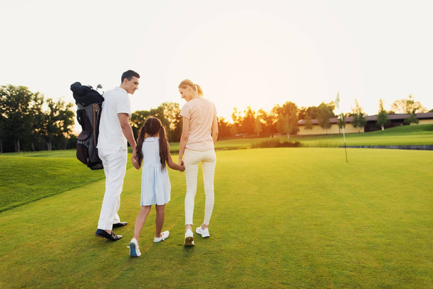 A man, a girl and a woman are walking along the golf course towards the sunset, holding hands - Foto, imagen