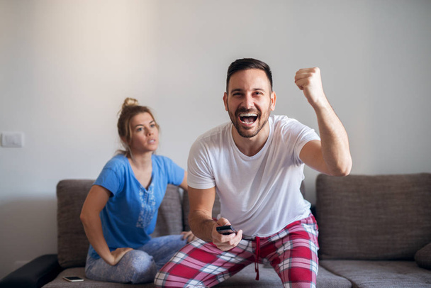 uśmiechnięty mężczyzna obchodzi wyniki sportowe w domu telewizor zdalnego przed denerwować dziewczynę podekscytowany - Zdjęcie, obraz