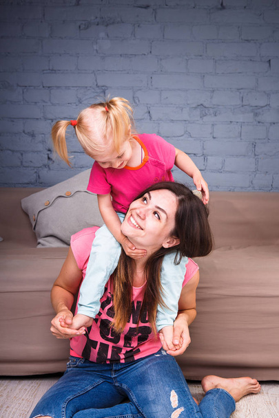 une jeune belle mère mince avec sa fille blonde sur ses épaules jouer, profiter et se livrer à des équipes près du canapé sur le sol en bois
 - Photo, image