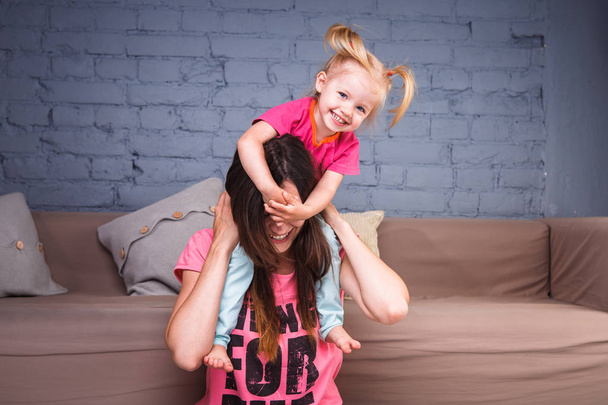 una joven y hermosa madre esbelta con su hija rubia sobre sus hombros jugar, disfrutar y disfrutar de los equipos cerca del sofá en el suelo de madera
 - Foto, Imagen