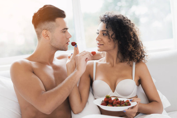 The guy and the girl are sitting on the bed in their underwear. They sexually feed each other with strawberries. - Foto, afbeelding