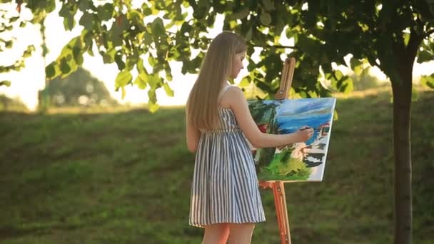 Hermosa chica dibuja un cuadro en el parque usando una paleta con pinturas y una espátula. caballete y lienzo con una imagen. El verano es un día soleado, puesta de sol
. - Metraje, vídeo