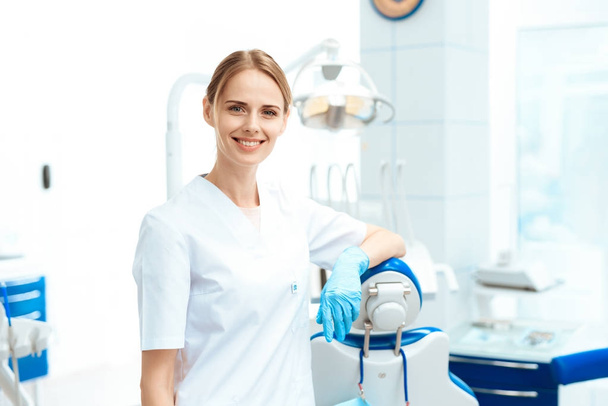 Female dentist posing against a background of dental equipment in a dental clinic. She puts on gloves - Foto, afbeelding