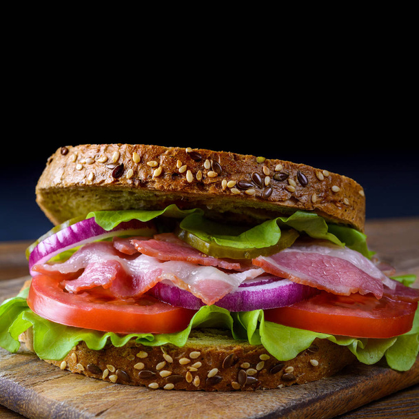 Sandwich recién preparado con tocino, tomate, pepino, cebolla sobre tabla de cortar de madera
 - Foto, imagen