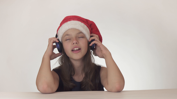 Güzel yaramaz kız genç bir Noel Baba şapkalı kulaklıklar müzik dinler ve boyunca üzerinde beyaz arka plan stok görüntüleri video şarkı söylüyor. - Video, Çekim
