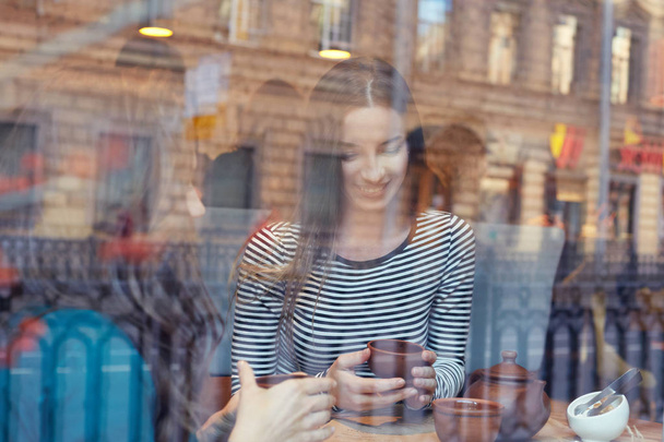 Zwei Studentinnen lächeln und lachen, während sie sich in der Mittagspause in der Mensa der Universität entspannen. Aufnahme attraktiver, gut gelaunter Damen, die durch das große Fenster eines modernen Cafés oder Restaurants Tee trinken. - Foto, Bild
