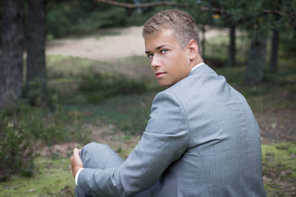 Gros plan extérieur de confiant jeune homme caucasien sérieux portant gris élégant costume formel relaxant, assis sur un banc dans la forêt. bel homme blond regardant la caméra à travers son épaule
 - Photo, image
