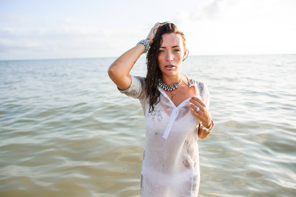 Sexy and beautiful wet brunette in long white shirt and bright jewelry staying in sea. Fashion photoshooting on the beach. - 写真・画像