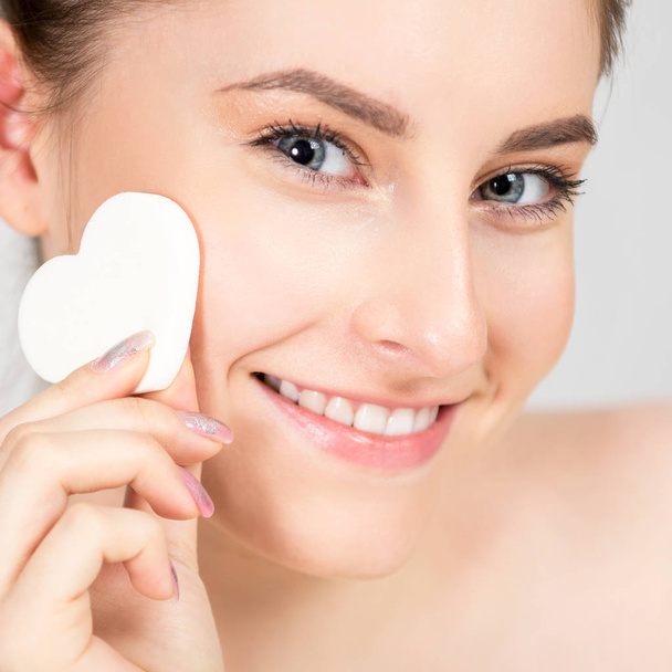 Portrait of a beautiful cheerful young woman with a sponge in the form of a heart in her hands. Her skin is fresh and healthy, and make-up is very light and natural. Photo closeup - Foto, afbeelding