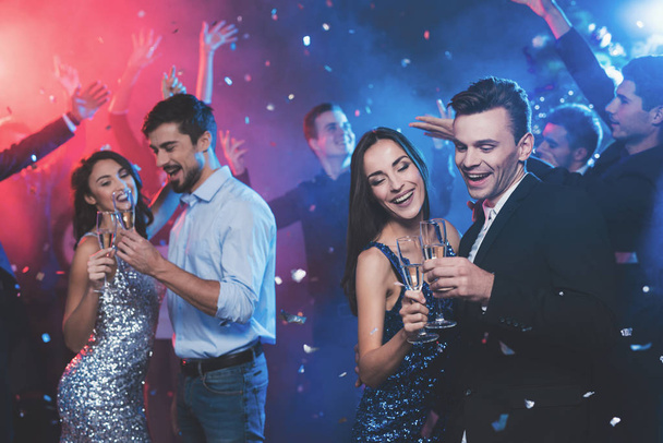 Young people have fun at a New Year's party. In the foreground two pairs dance. The girls are wearing gray and blue dresses in sequins. On one of the guys a light shirt, on the other a black jacket. - Foto, imagen