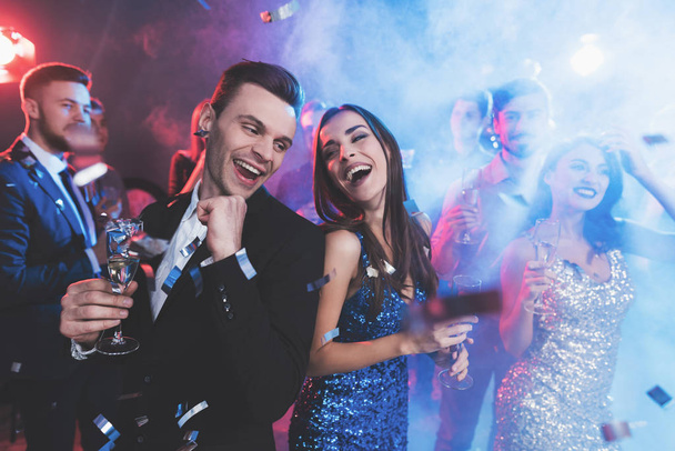 New Year party. Young couple dancing with glasses of champagne in hands. Around fly confetti and there is white smoke. - Фото, зображення