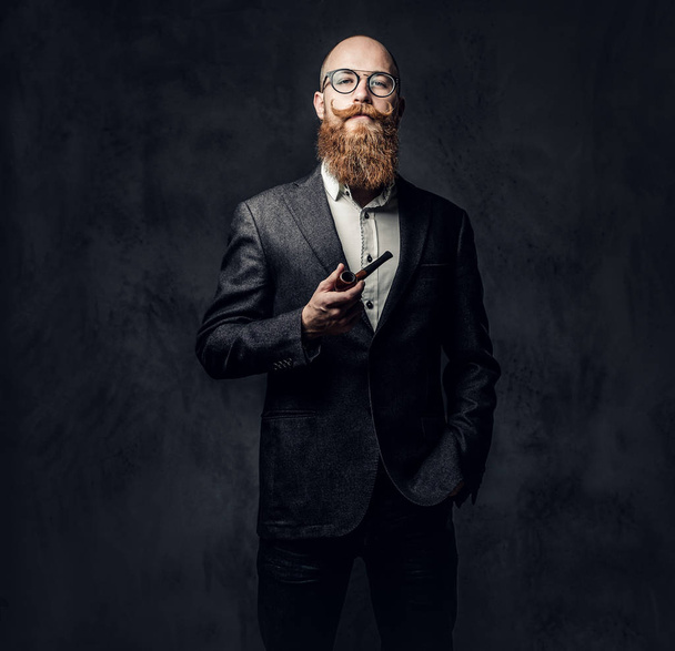 Redhead bearded male dressed in a suit and eyeglasses smoking tradition pipe over dark grey background. - Fotoğraf, Görsel