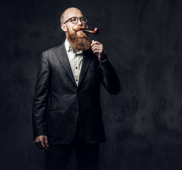 Pelirroja barbudo macho vestido con un traje y gafas de fumar tradición pipa sobre fondo gris oscuro
. - Foto, Imagen