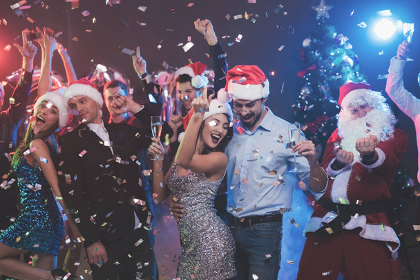 Young couple dancing with glasses of champagne in hands. Behind them dance their friends and man dressed as Santa Claus. - Fotó, kép