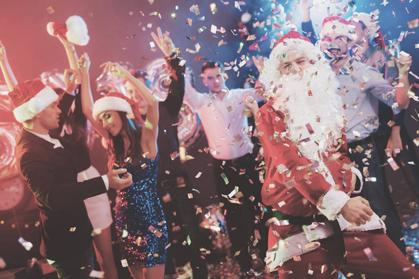 Un uomo vestito da Babbo Natale si diverte a una festa di Capodanno. Insieme a lui hanno amici divertenti. Intorno a loro mosche coriandoli, su uno sfondo di foschia multicolore. Le persone sono molto allegre
. - Foto, immagini