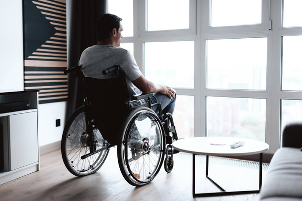 The disabled person in a wheelchair sits in front of a large panoramic window and looks into it. He is sad. He is in the living room of his modern apartment. - Foto, immagini