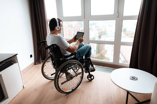 The disabled person in a wheelchair sits in front of a large panoramic window and looks at something on his tablet. - Photo, Image