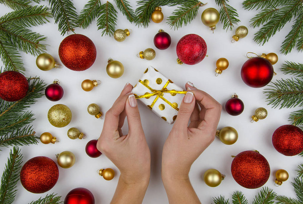 Weihnachtskomposition aus goldenen und roten Weihnachtskugeln, Zapfen und Tannenzweigen. auf weißem Hintergrund weibliche Hände, die eine kleine Geschenkschachtel halten. Winterflach gelegt - Foto, Bild