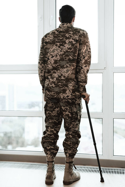 A disabled soldier is leaning on a crutch by the window. He looks into the large panoramic window. - Photo, Image