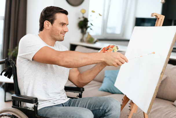 The disabled person is sitting in a wheelchair opposite the easel. He draws a picture. He is in his large bright living room. He smiles. - Фото, изображение