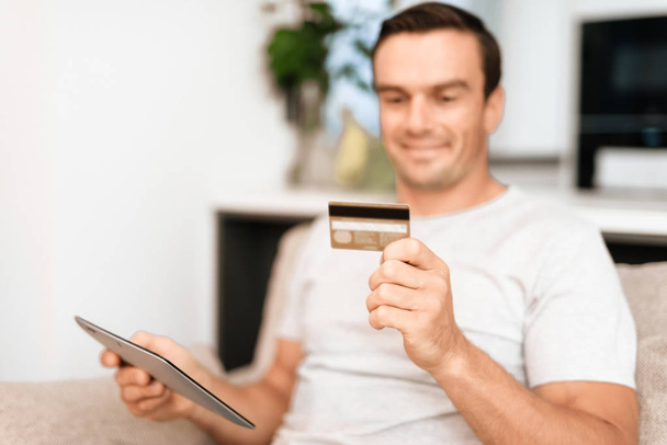 A man is sitting on the couch. He has a tablet and a credit card in his hands. He wants to buy something online. - Foto, Imagem