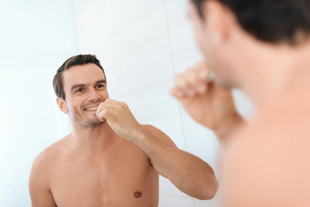 A man is standing in the bathroom in the morning and brushing his teeth. He smiles. He stands in a bright bathroom and looks at the gray in the mirror. - Foto, Imagen